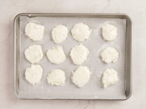 Twelve round portions of white cheese spread are arranged on a parchment-lined baking sheet placed on a light-colored surface.