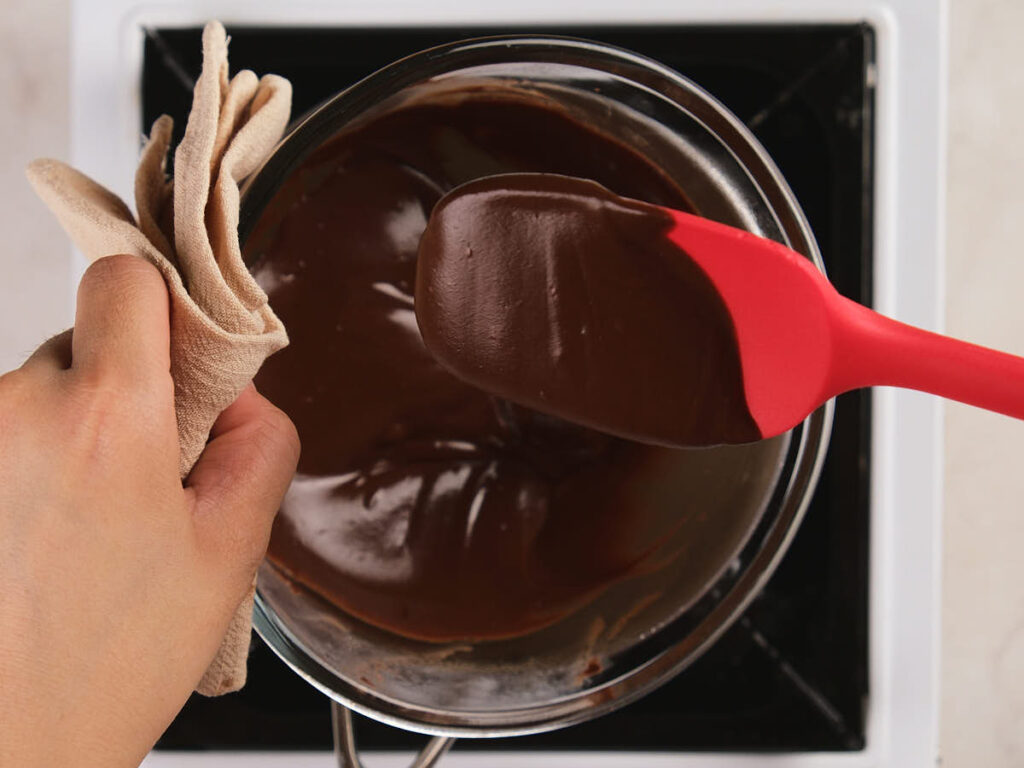 A hand holds a towel and stirs melted chocolate in a glass bowl over a pot on a stovetop using a red spatula.