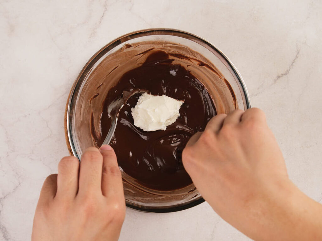 A person mixes a bowl of melted chocolate with a dollop of cream or butter on a light-colored countertop.