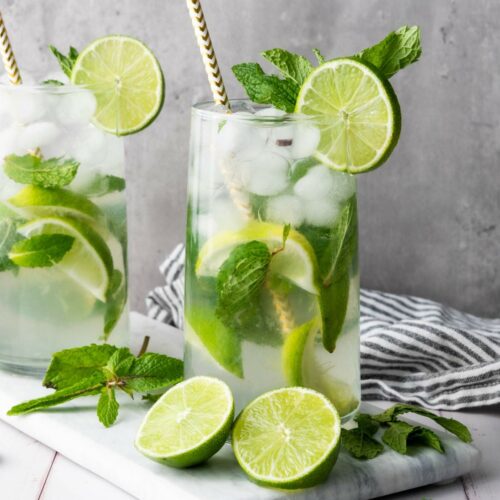 Two tall glasses of iced mojito with lime slices, mint leaves, and striped straws on a marble board.