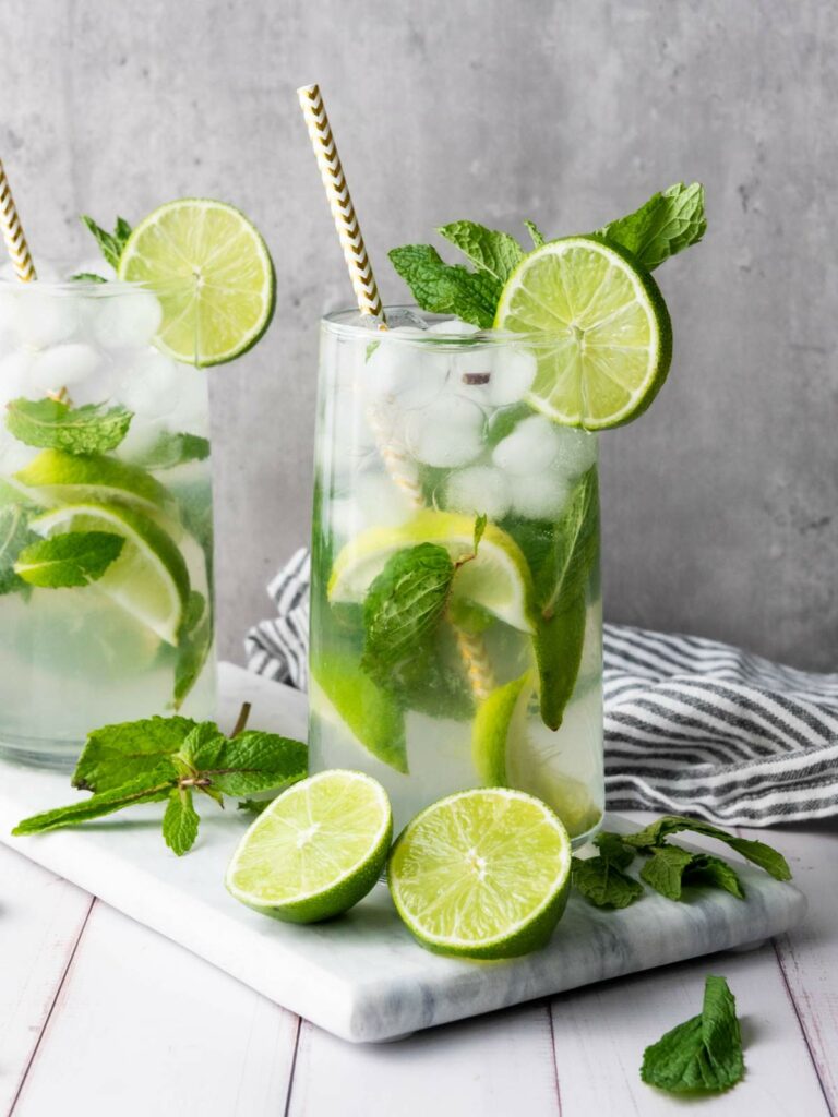 Two tall glasses of iced mojito with lime slices, mint leaves, and striped straws on a marble board.