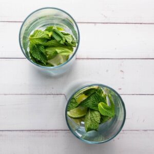 Two clear glasses on a white wooden surface, each containing mint leaves and lime wedges.