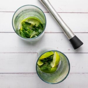 Two glasses with lime wedges and mint leaves, accompanied by a metal muddler, sit on a white wooden surface.