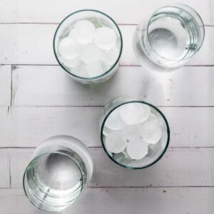 Four glasses of water are placed on a white wooden surface; two glasses contain ice cubes, while the other two hold only water.
