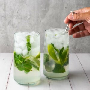Two glasses of mojito with ice, mint leaves, and lime wedges on a white table.