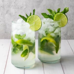 Two glasses of mojito with ice, lime slices, and fresh mint on a white table against a light gray background.