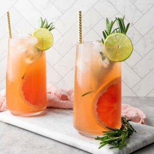 Two tall glasses of iced grapefruit cocktail garnished with lime slices and rosemary sprigs, placed on a marble board with a pink cloth in the background.