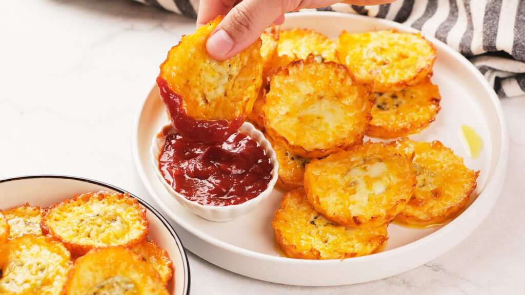 A hand dips a cheesy, baked pickle chips into a small bowl of red sauce on a white plate.