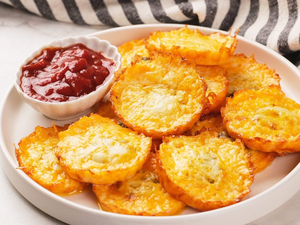 A plate of baked cheesy pickle chips with a small bowl of red dipping sauce, set on a white surface with a striped cloth in the background.