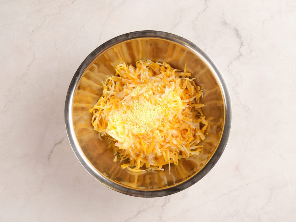 A stainless steel mixing bowl containing shredded cheese and seasoning on a light marble surface.