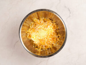A stainless steel mixing bowl containing shredded cheese and seasoning on a light marble surface.