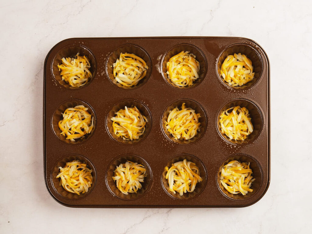 A muffin tin with twelve cups, each filled with a mixture of shredded yellow and white cheese, sits on a white surface.