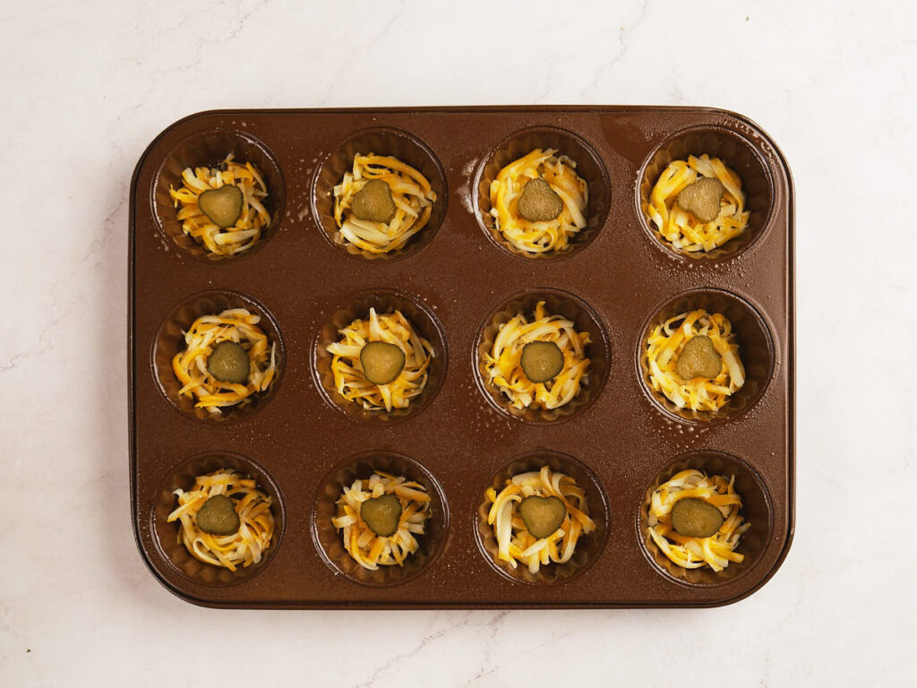 A muffin tin with twelve cups, each filled with shredded cheese and topped with a round pickle slice, on a white surface.