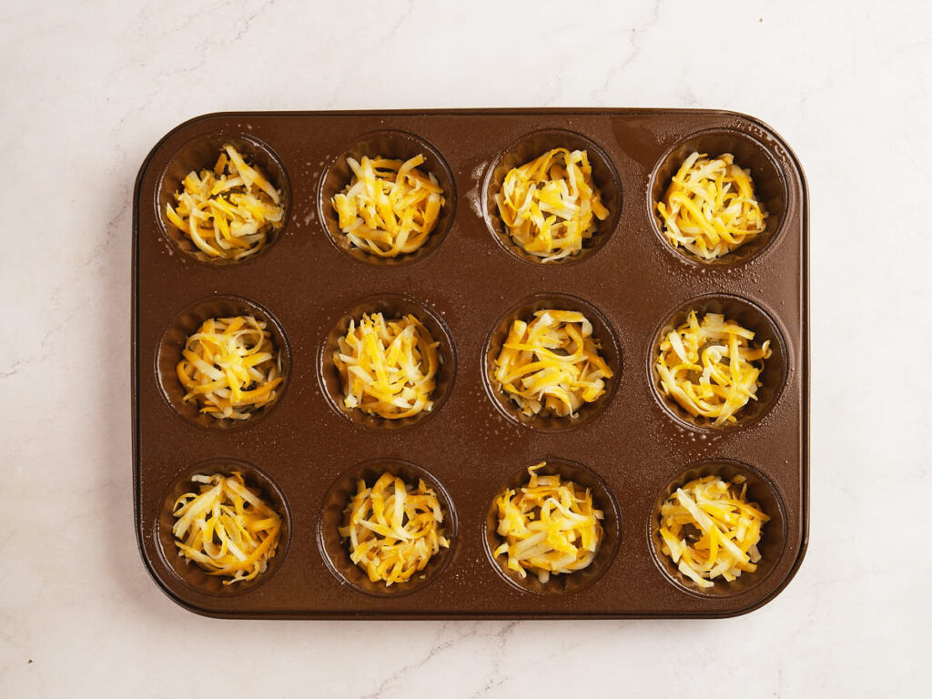 A muffin tin with twelve cups, each filled with shredded cheese, sits on a light-colored marble surface.