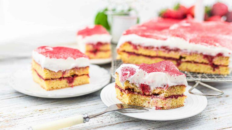 Several slices of layered sponge cake with red jam and white frosting—perfect sugar-free desserts—are displayed on plates and a cooling rack, with a fork on one plate.