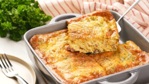 A baked omelet with cheese and herbs is being served from a square baking dish with a spatula.