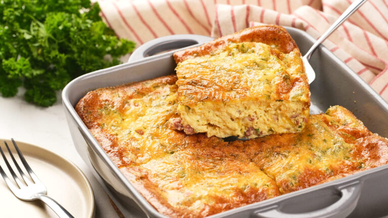 A baked omelet with cheese and herbs is being served from a square baking dish with a spatula.