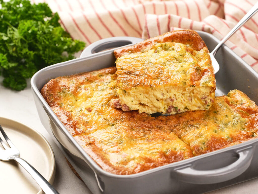 A baked egg casserole with a golden top is being served from a square baking dish.