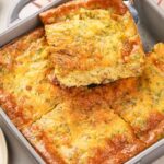 A baking dish with a golden-brown egg casserole, cut into squares, with one piece being lifted out.