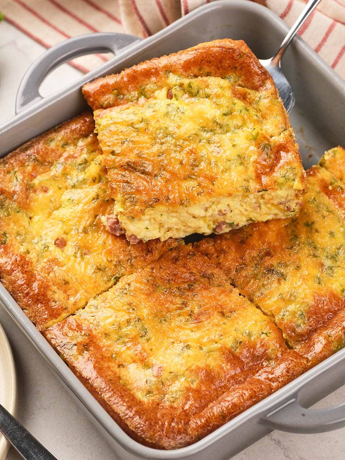 A baking dish with a golden-brown egg casserole, cut into squares, with one piece being lifted out.