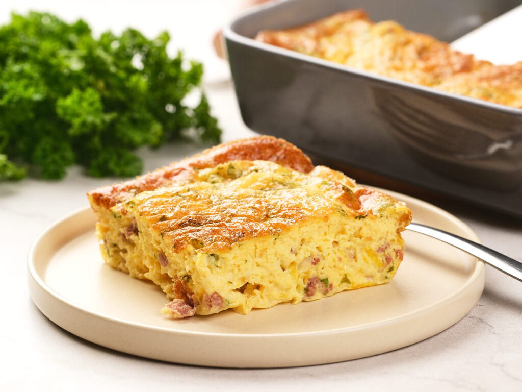 A slice of baked omelet on a beige plate, with a baking dish and fresh parsley in the background.