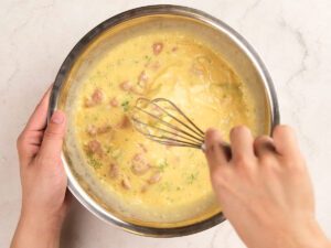 A person whisking eggs with chopped green vegetables and meat pieces in a metal bowl on a light countertop.