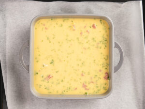 A square baking pan filled with an uncooked egg mixture containing herbs and chopped ingredients.