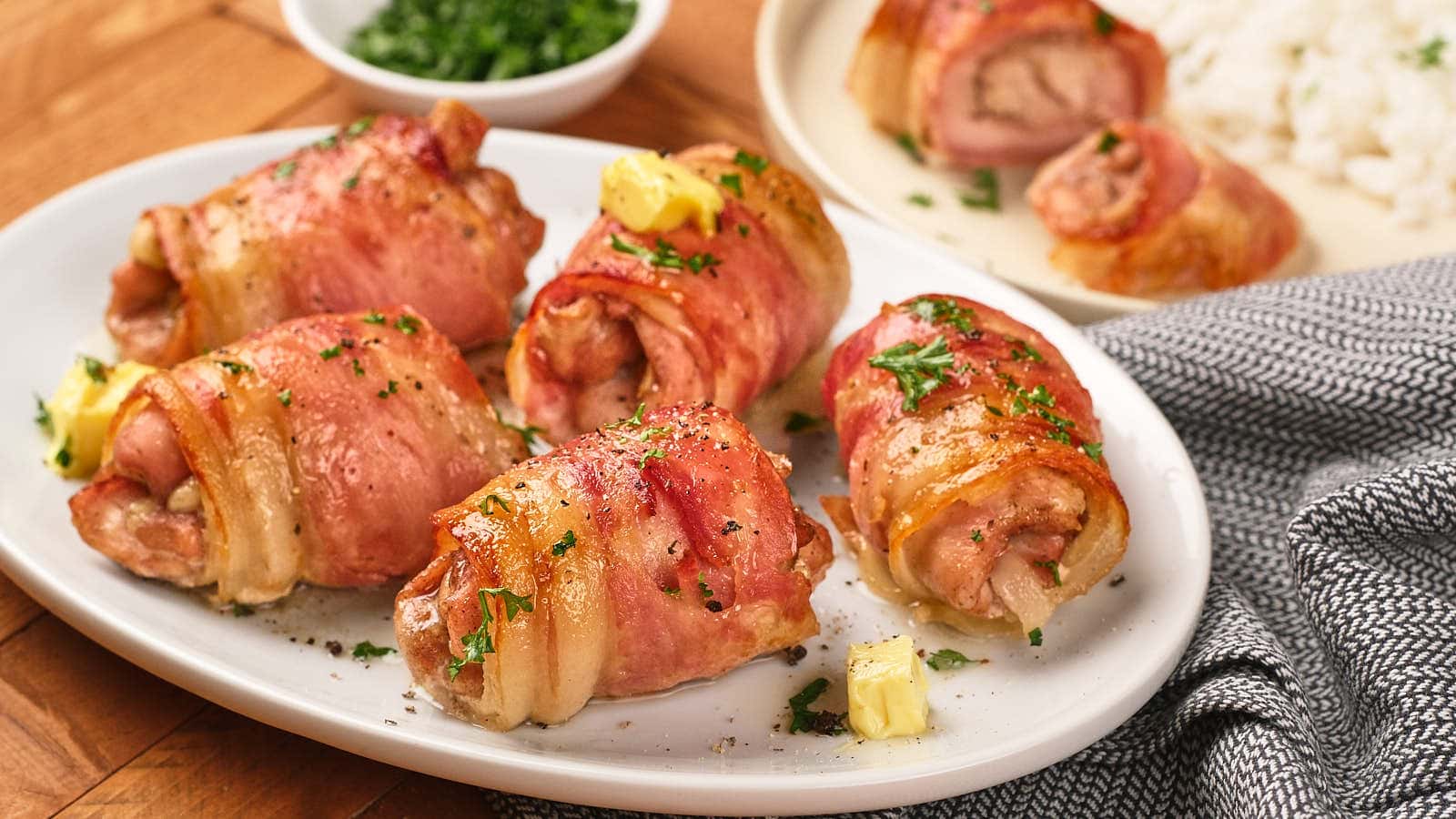 Six pieces of bacon-wrapped chicken garnished with herbs and small butter cubes.