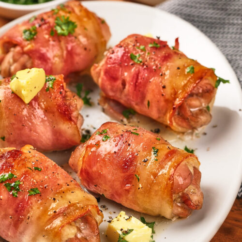 Five bacon-wrapped chicken thighs garnished with parsley and small pieces of butter are arranged on a white plate.