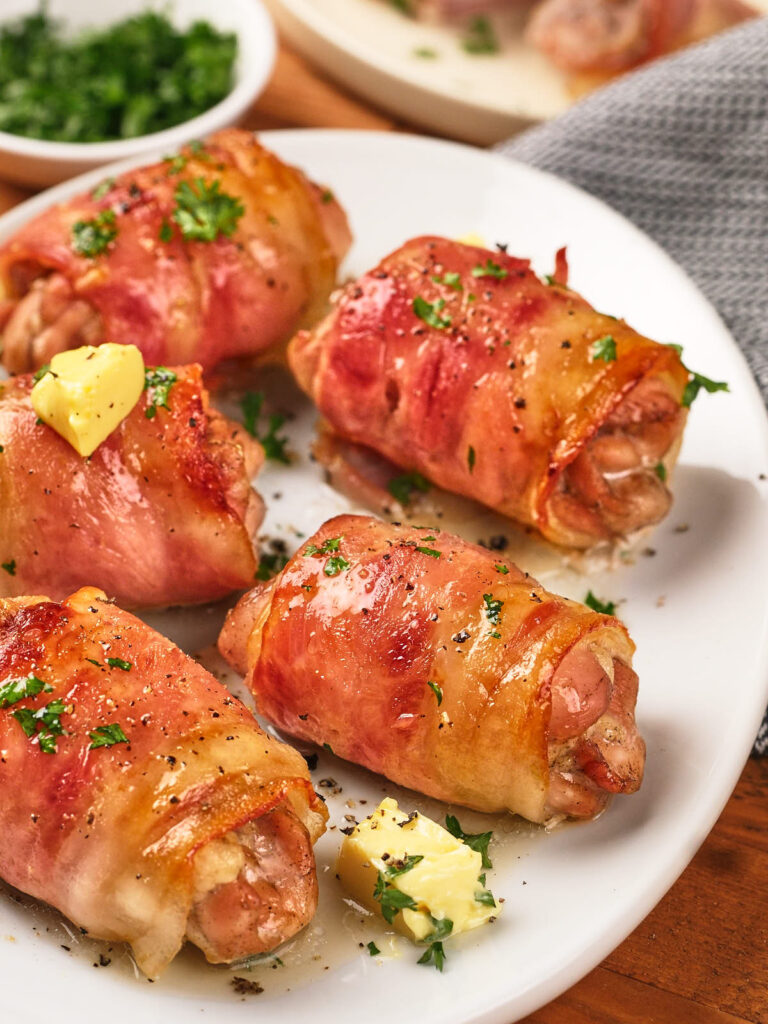 Five bacon-wrapped chicken thighs garnished with parsley and small pieces of butter are arranged on a white plate.