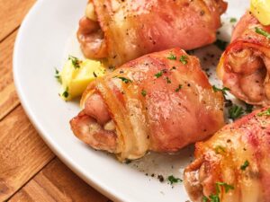 Close-up of bacon-wrapped chicken thighs on a white plate, garnished with herbs and served with wedges of butter.