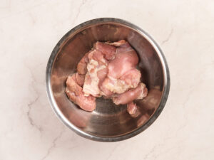 Raw pieces of chicken placed in a stainless steel mixing bowl on a light-colored marble countertop.