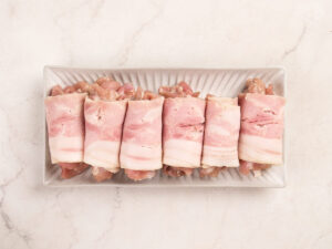 Six pieces of raw meat wrapped in bacon are neatly arranged in a rectangular white dish on a light marble surface.