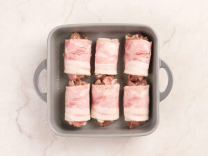 A baking dish with six pieces of raw, bacon-wrapped meat arranged in two rows on a light countertop.