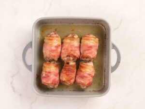 A square baking dish with six pieces of bacon-wrapped chicken, cooked and arranged in two rows on a light-colored countertop.