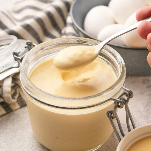 A spoon scoops creamy egg pudding from a glass jar, with eggs and a striped cloth in the background.