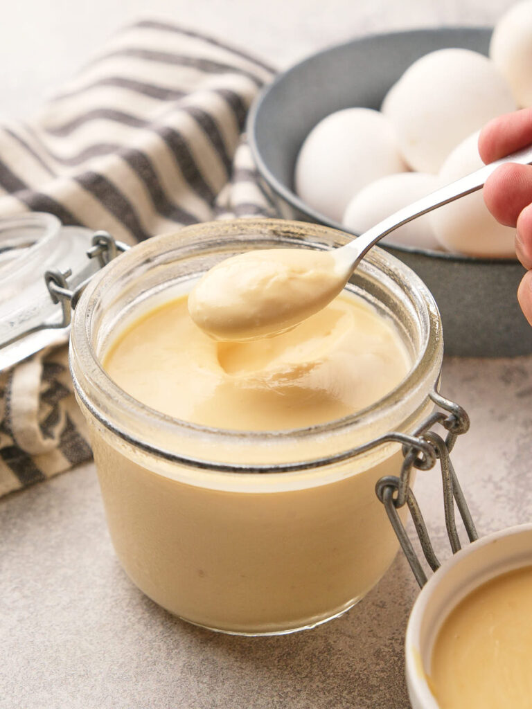 A spoon scoops creamy egg pudding from a glass jar, with eggs and a striped cloth in the background.