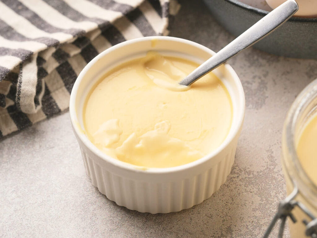 A small white ramekin filled with egg pudding, with a spoon resting inside, sits on a light countertop next to a striped cloth.