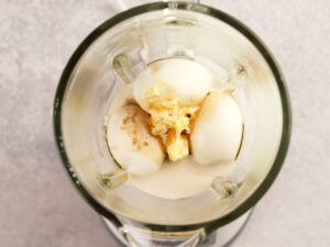 Overhead view of a blender containing eggs, oil, garlic, and seasonings on a light countertop.