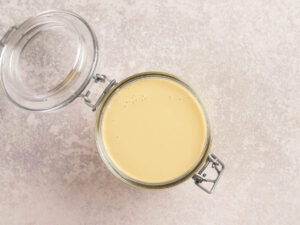 A glass jar filled with creamy yellow liquid, viewed from above, with the lid open on a light-colored textured surface.