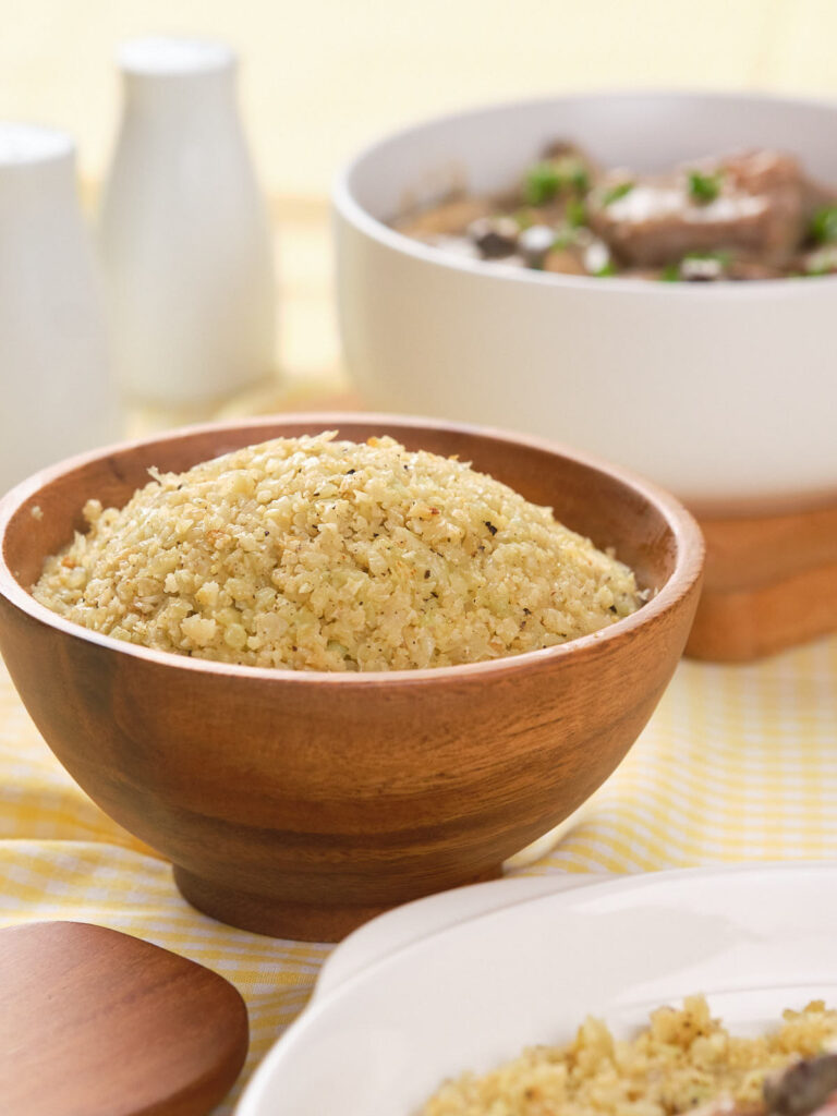 A wooden bowl filled with seasoned couscous sits on a yellow checkered cloth, with a white serving bowl and white containers in the background.