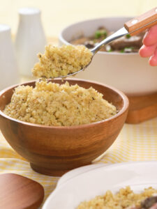 A hand holds a spoonful of cooked cauliflower rice above a wooden bowl filled with more cauliflower rice, with dishes and condiments in the background.