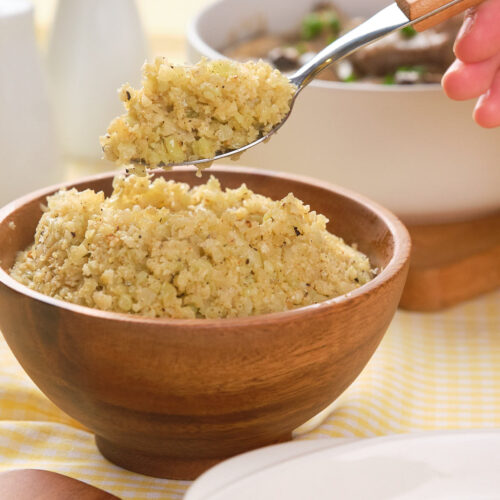A hand holds a spoonful of cooked cauliflower rice above a wooden bowl filled with more cauliflower rice, with dishes and condiments in the background.