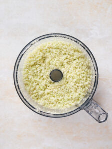 Overhead view of chopped cauliflower pieces in a food processor on a light-colored surface.