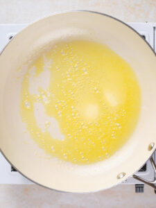 A white skillet on a stovetop containing melted butter that is starting to bubble and foam.