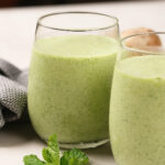 Two glasses filled with a green smoothie sit on a light surface, with a sprig of fresh mint placed in front of them.