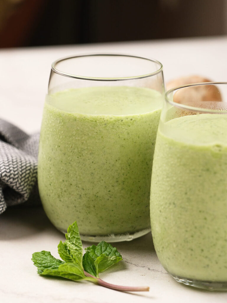 Two glasses filled with a green smoothie sit on a light surface, with a sprig of fresh mint placed in front of them.