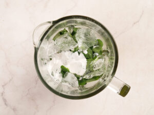 A blender pitcher viewed from above, containing ice cubes, spinach leaves, and a small amount of white liquid on a light marble surface.