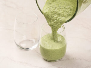 A thick green smoothie is being poured from a blender into a clear glass, with an empty glass beside it on a light-colored surface.