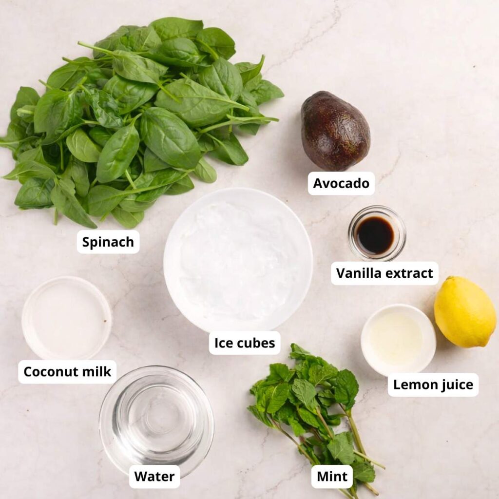 Ingredients for a smoothie laid out on a white surface: spinach, avocado, mint, ice cubes, coconut milk, water, lemon juice, and vanilla extract.